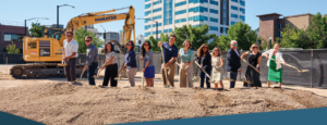 Group of professionals at a groundbreaking ceremony with construction equipment and city buildings in the background for a five-year capital improvement plan.
